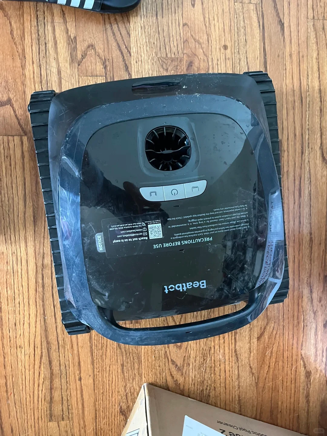 Top-down shot of a black Beatbot cordless robotic pool cleaner resting on a hardwood floor, showing its tank tracks, control buttons, and handle.