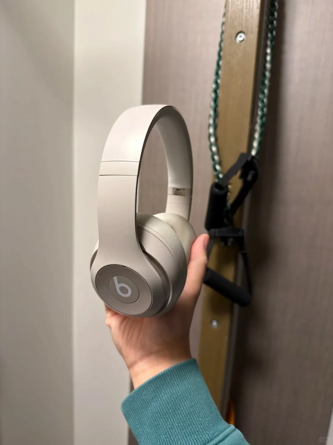A hand holds a pair of light beige Beats over-ear headphones indoors against a wall.