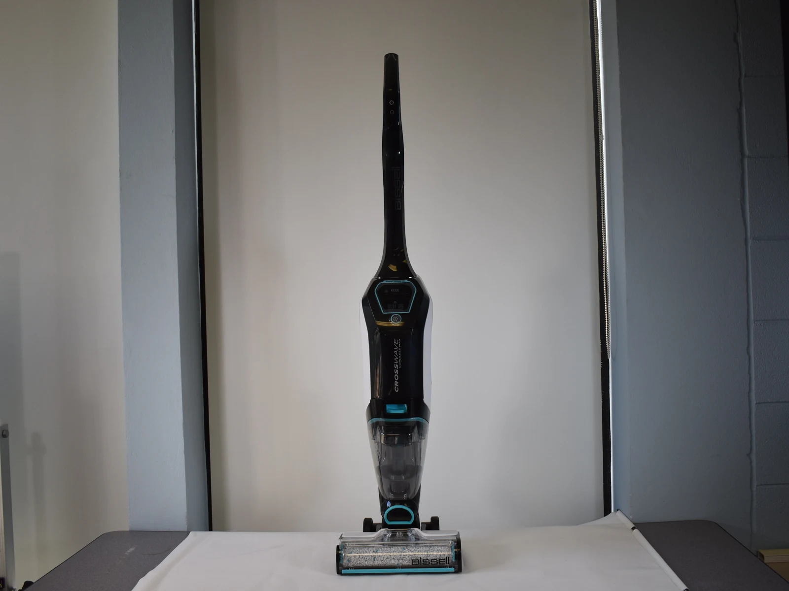 Front-facing black BISSELL CrossWave Cordless Max wet-dry floor cleaner standing upright on a white cloth, with a transparent tank, digital display, wheels, and roller head visible.