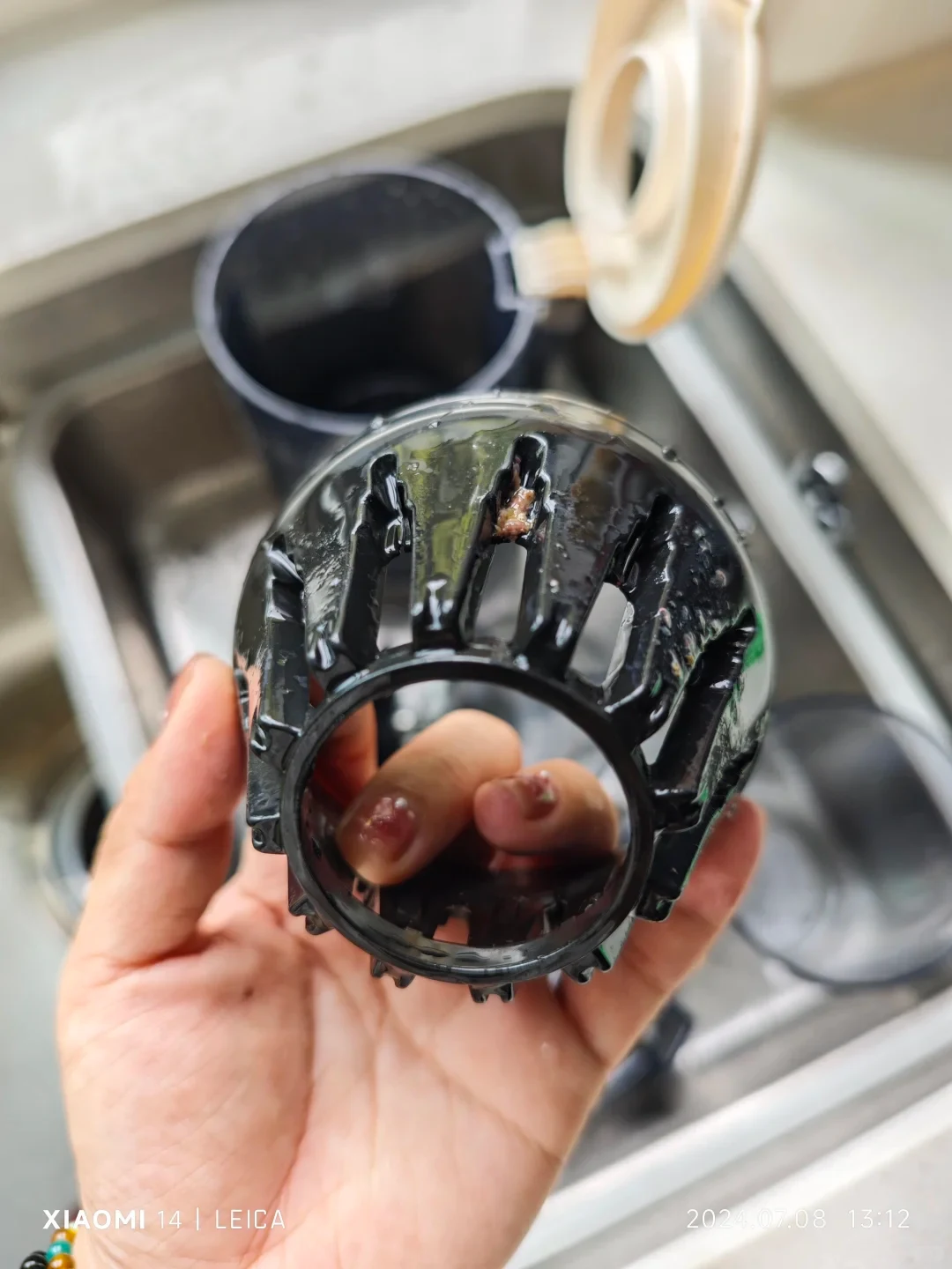 A hand holds the black slotted juicer filter component over a kitchen sink, with wet pulp still lodged in its ribs and the open hopper behind it.