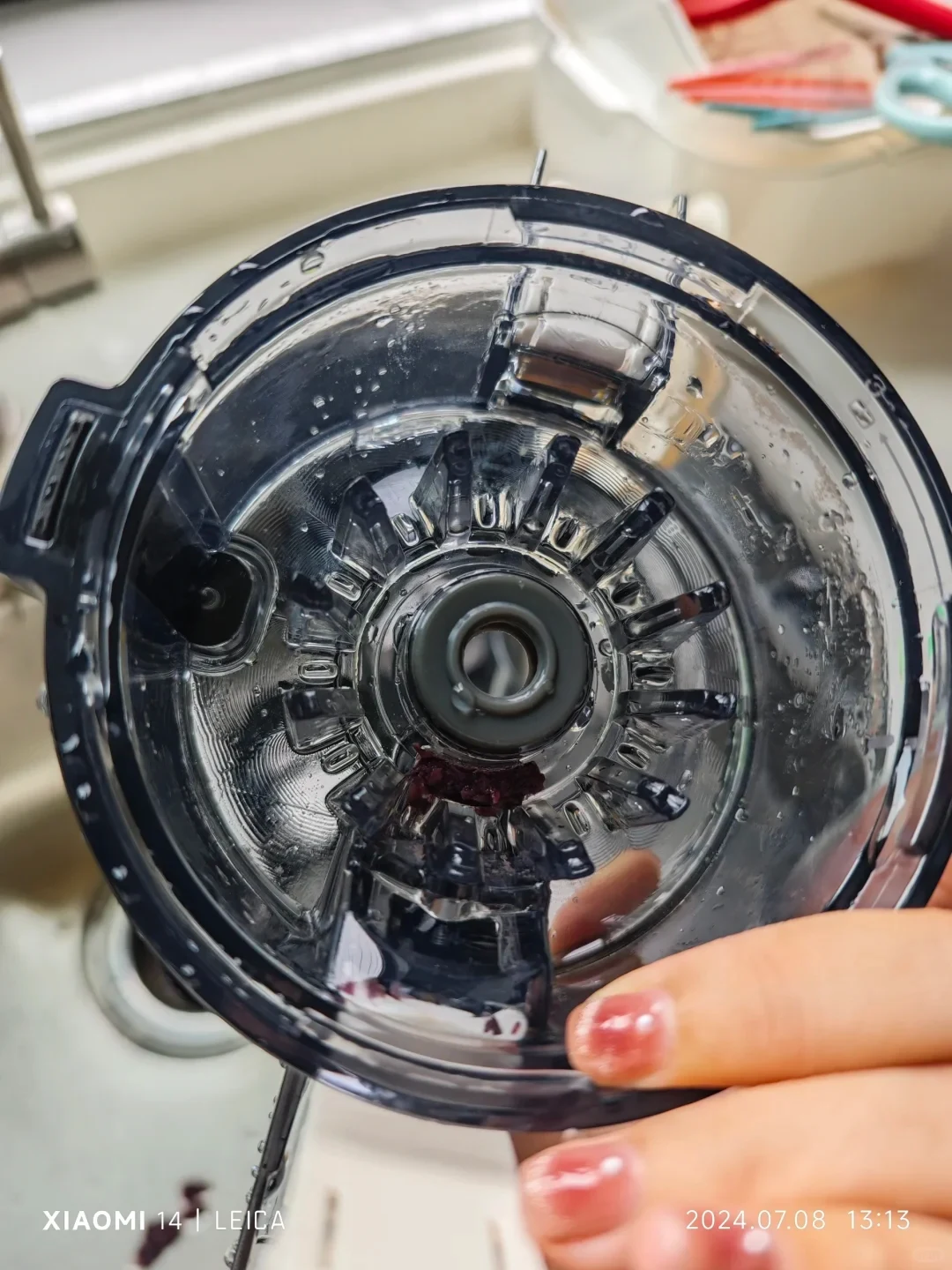 A hand holds the underside of the clear juicer chamber over the sink, showing droplets and residue around the central opening and radial grooves.