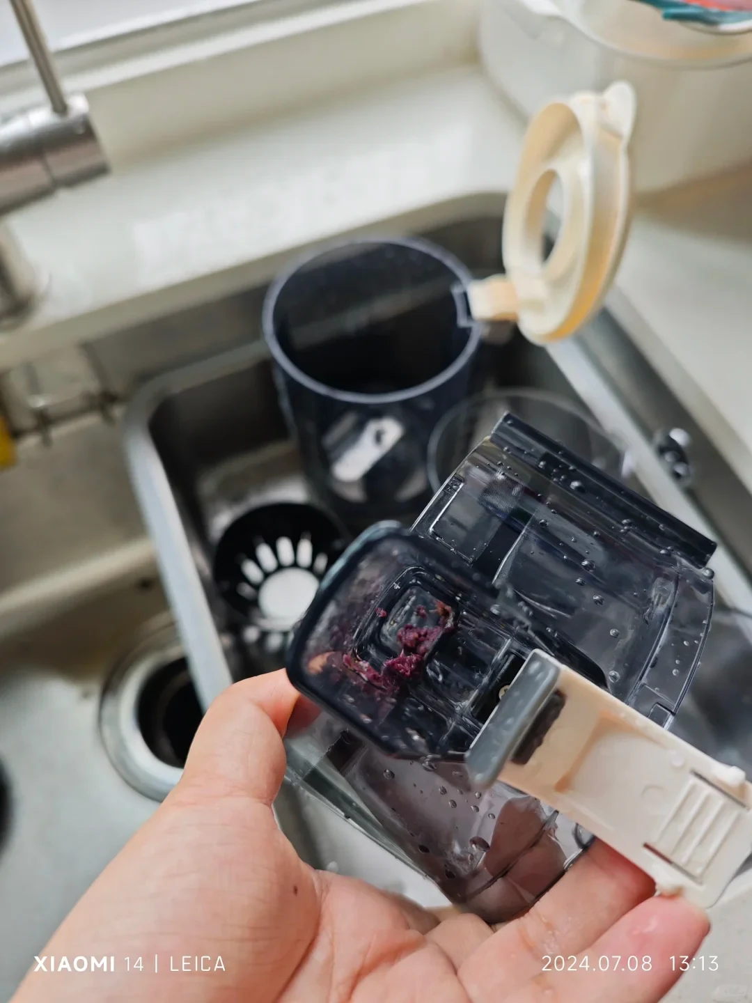 A hand holds the clear juicer chamber sideways, showing reddish pulp stuck by the small outlet and beige outlet flap; other juicer parts rest in the sink.