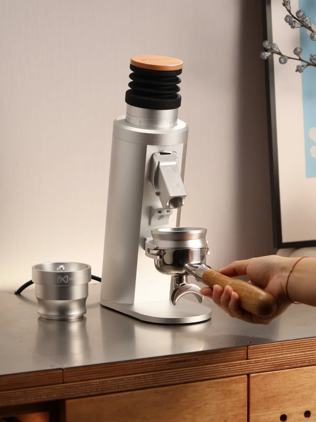 A hand holds a wooden-handled portafilter beneath a silver espresso grinder, with a separate metal cup on the counter beside it.