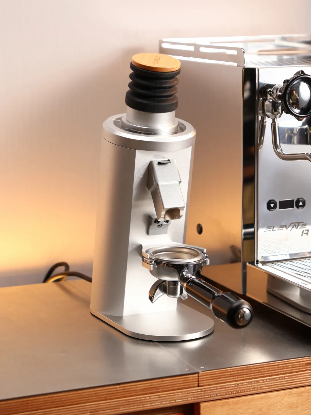 A silver coffee grinder with a wooden-topped black bellows stands beside a stainless-steel espresso machine. A portafilter is positioned beneath the grinder’s chute on the counter.
