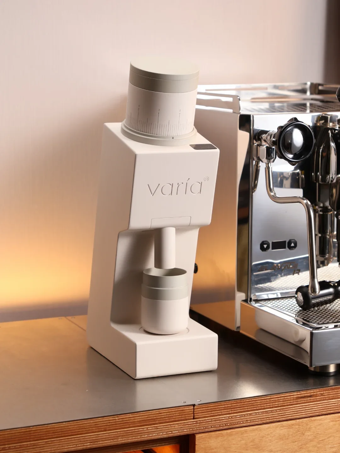 A white Varia coffee grinder stands beside a stainless-steel espresso machine on a counter. Its cylindrical adjustment dial and grounds cup are visible.