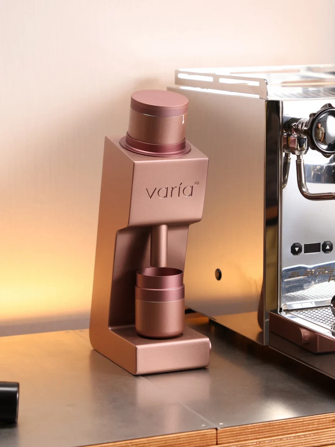 A rose-gold Varia coffee tool stands on a counter beside a stainless-steel espresso machine.