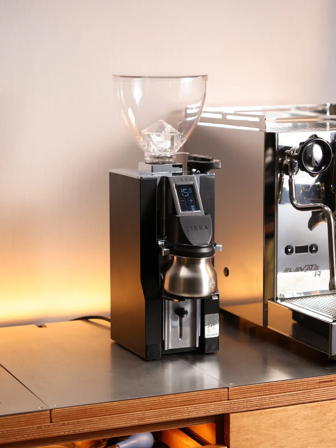 A black LIBRA coffee grinder with a clear bean hopper and metal grounds container sits on a counter beside a stainless-steel espresso machine.