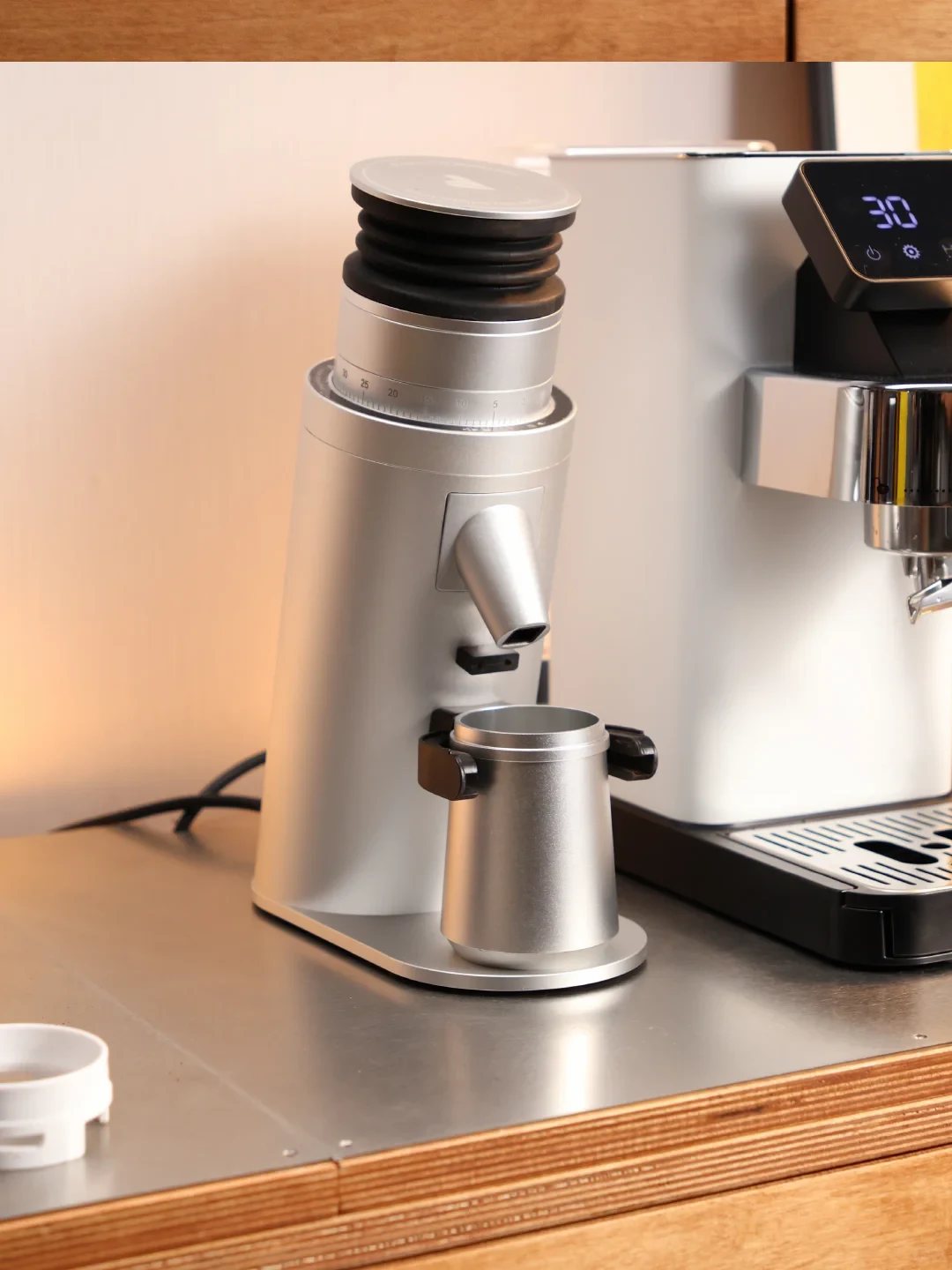 A silver coffee grinder with a stepped black bellows top and numbered adjustment ring sits beside an espresso machine on a counter; its display reads 38.