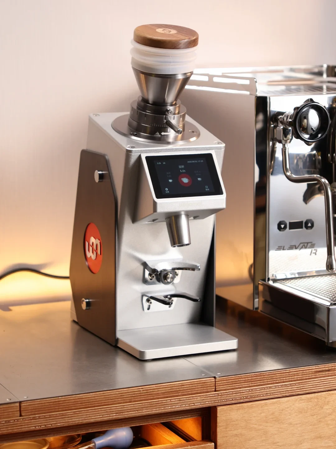 A stainless-steel coffee grinder with a wooden lid and touchscreen stands beside a stainless-steel espresso machine on a counter. The grinder’s screen shows a double-cup setting and a 5.0-second value.