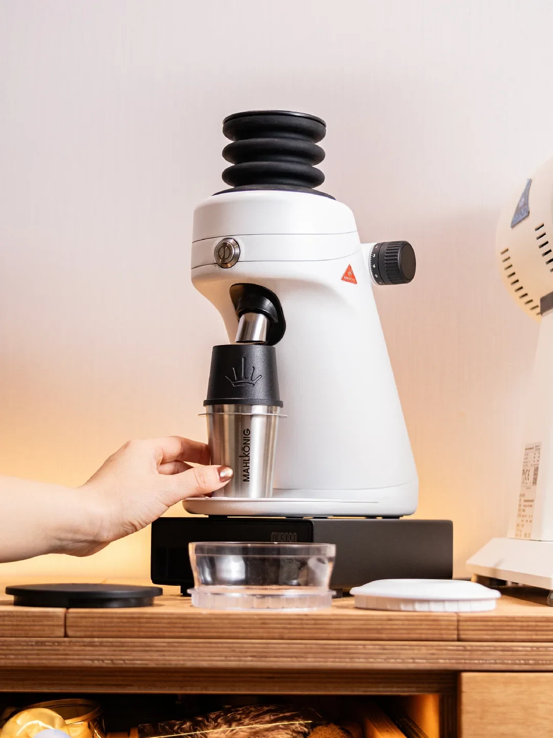 A white coffee grinder stands on a black base on a wooden counter. A hand holds a stainless-steel cup beneath its dispensing spout, with a clear cup and several lids nearby.