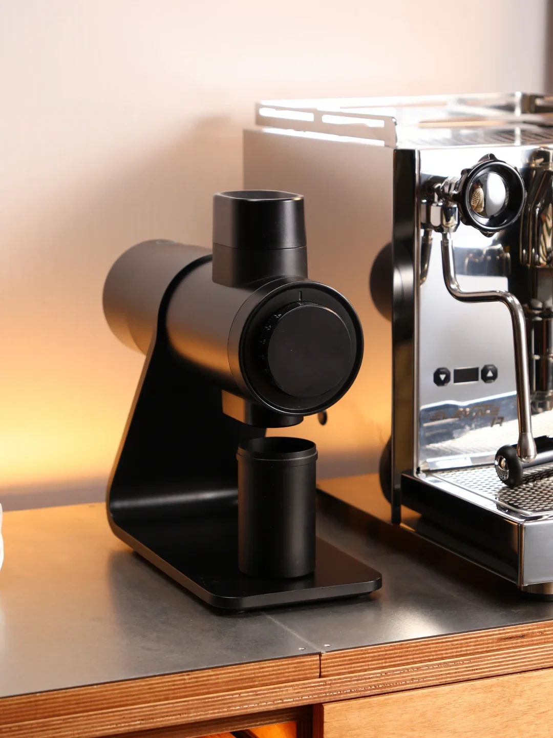 A black coffee grinder with a cylindrical grounds container sits on a counter beside a stainless-steel espresso machine.