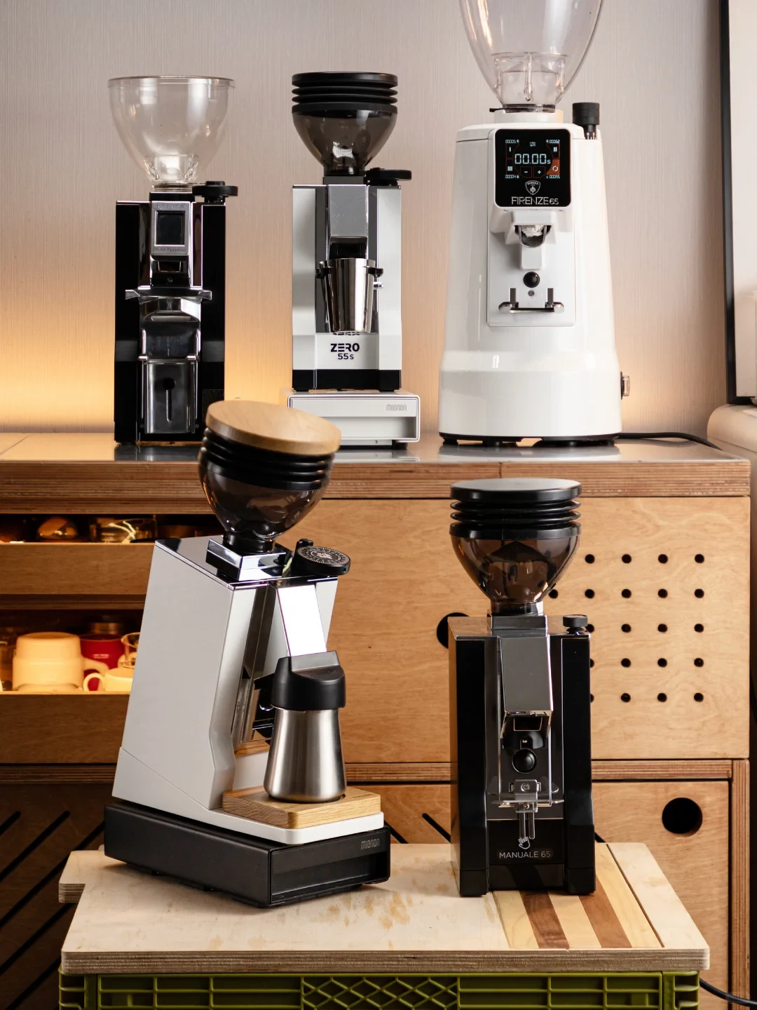 Several coffee grinders in black, silver, and white are displayed on wooden counters and a small table; some have bean hoppers, and the white grinder has a digital display.