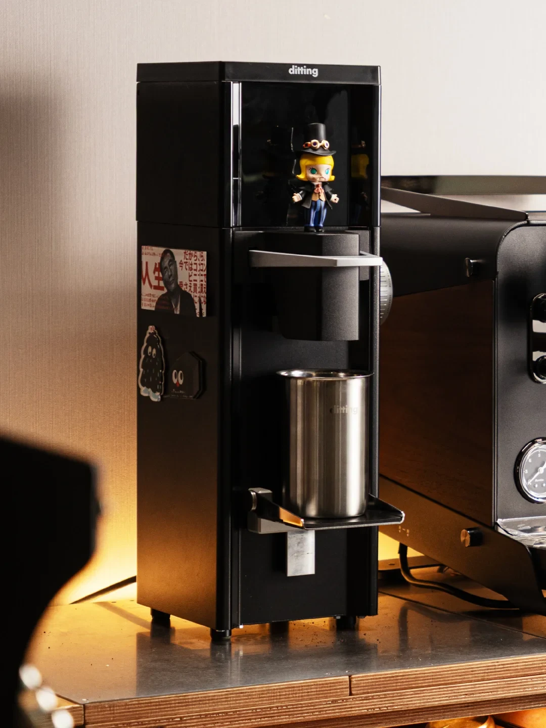 A black Ditting grinder stands on a counter beside an espresso machine, with a metal grounds cup below the chute, decorative stickers on the side and a figurine on its upper shelf.