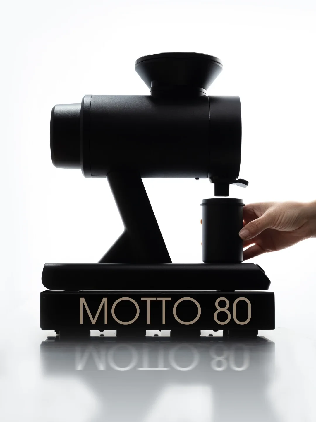 A black MOTTO 80 machine is shown against a white background as a hand holds a cylindrical container beneath its outlet; the product name is reflected on the surface below.