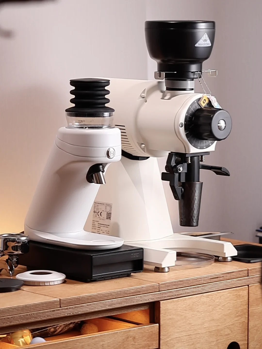 Two white coffee grinders sit on a wooden counter, with a black hopper mounted on the larger grinder and a black digital scale beneath the smaller one.