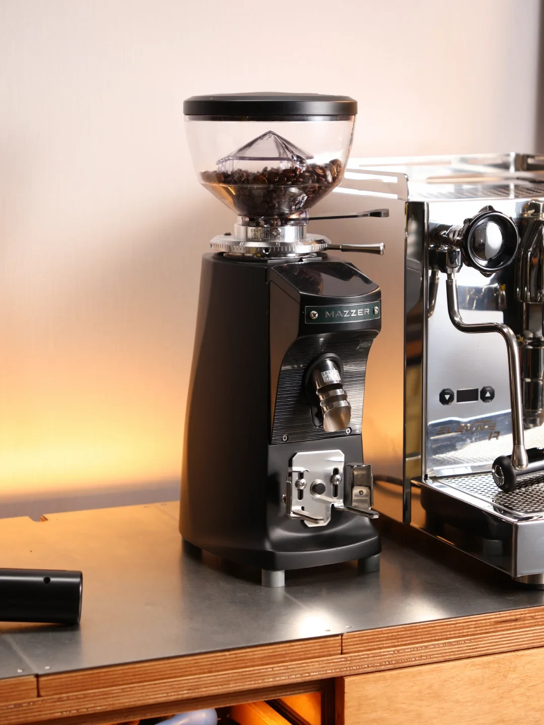 A black Mazzer coffee grinder with a clear hopper of coffee beans stands beside a stainless-steel espresso machine on a counter.