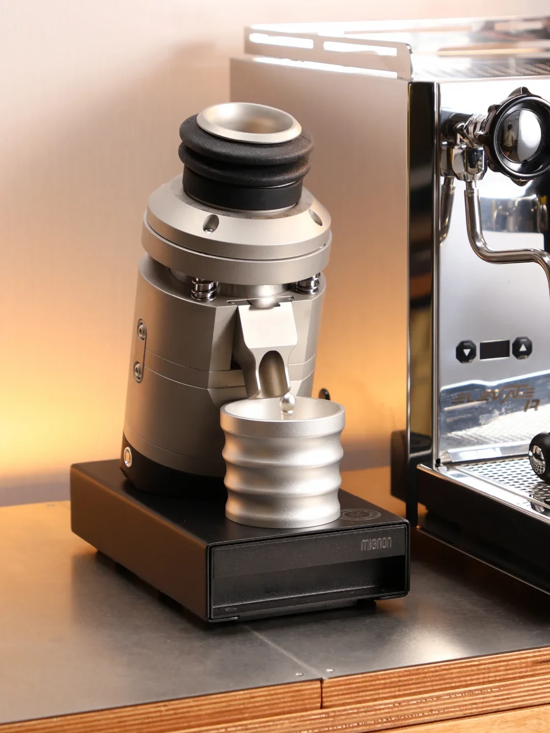A metallic coffee grinder with a black base and ribbed silver grounds container sits beside a stainless-steel espresso machine on a counter.