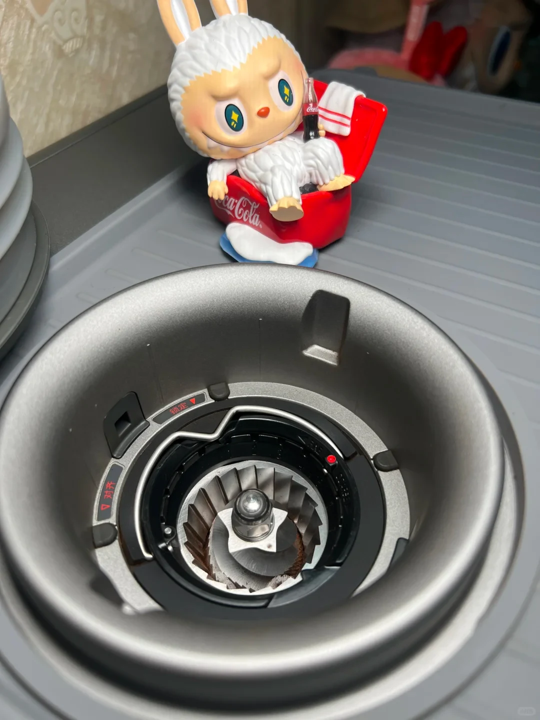 A close-up view looking down into the conical burr chamber of an electric coffee grinder, with a Labubu figurine seated in a red Coca-Cola cooler tub resting on a gray mat in the background.