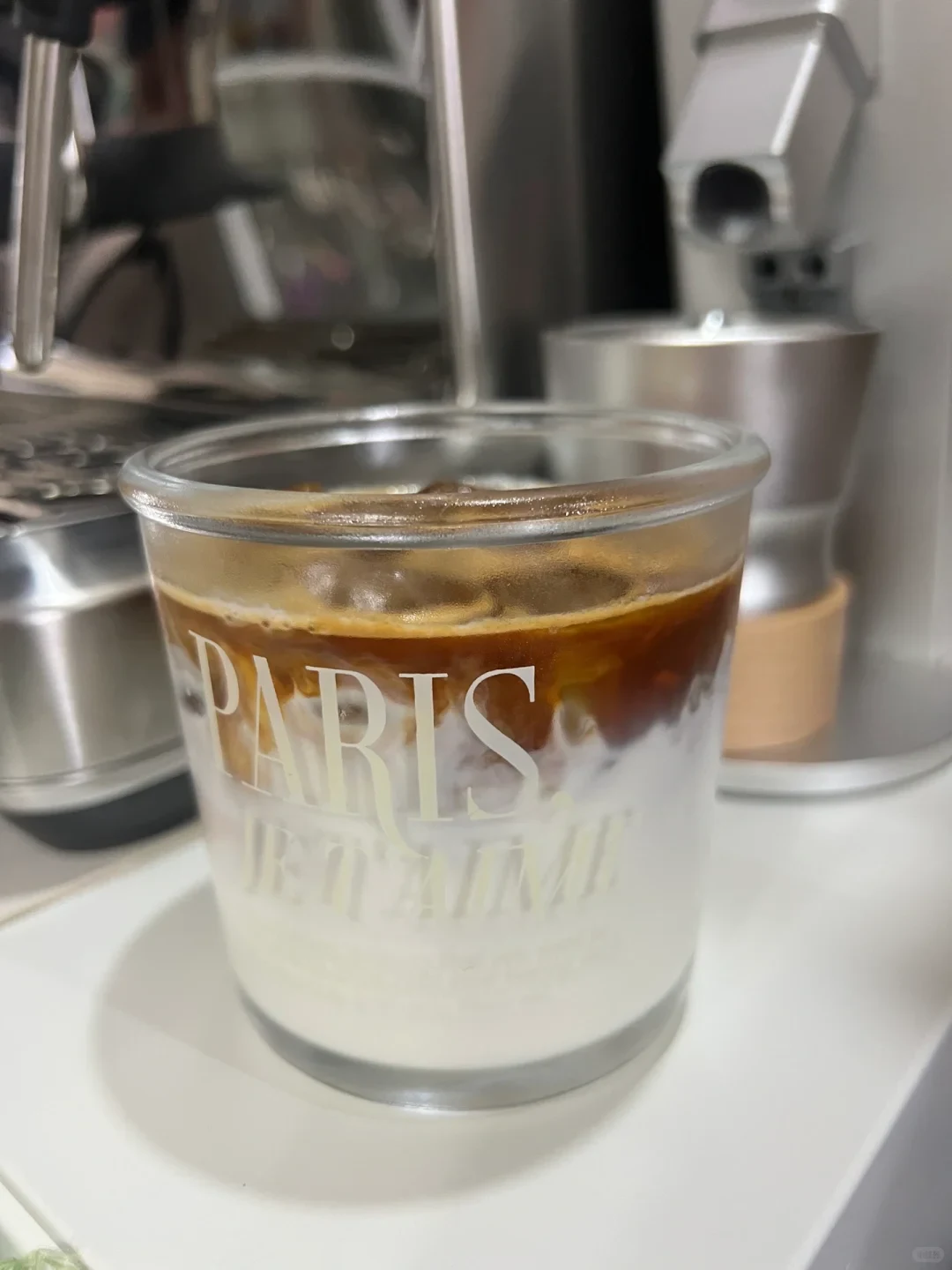 A glass cup containing an iced dirty latte sits on a counter in front of an espresso machine setup. The drink has dark brown espresso layered over chilled white milk with ice, and the glass is printed with the words 'PARIS JE T'AIME'.