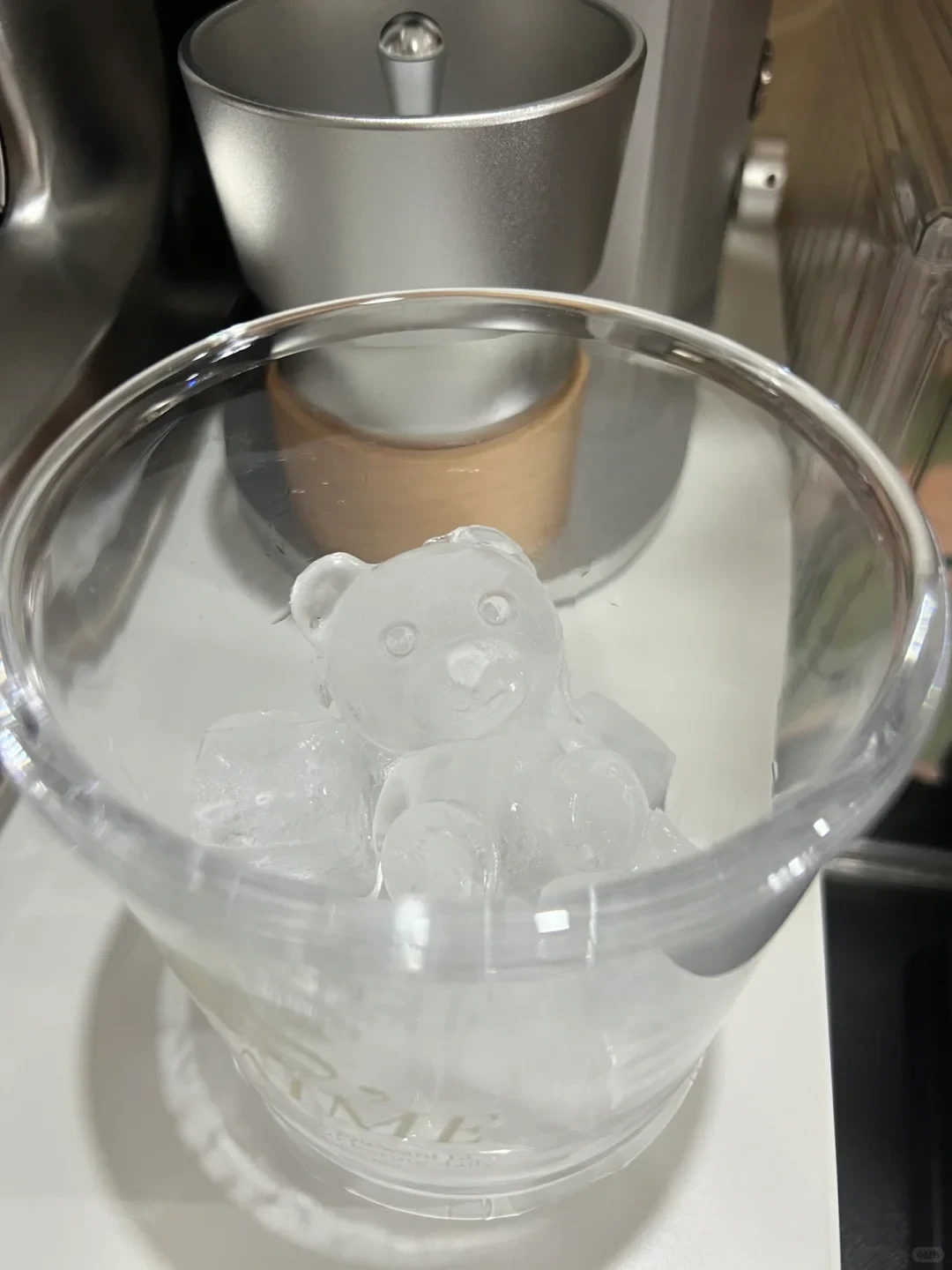 A clear glass tumbler holding clear ice, including an ice sculpture shaped like a small teddy bear, set in front of a silver coffee appliance.