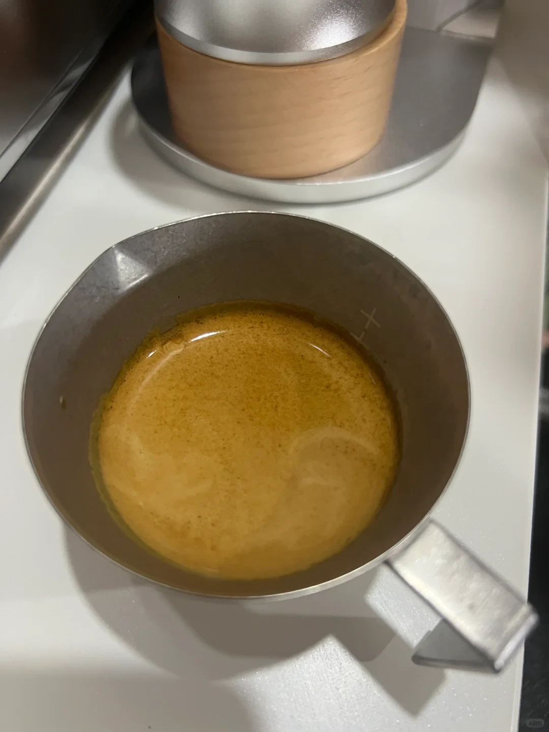 A top-down view of a matte metal espresso measuring cup filled with freshly brewed espresso covered in a rich, golden-brown layer of crema, resting on a white countertop next to coffee-making equipment.