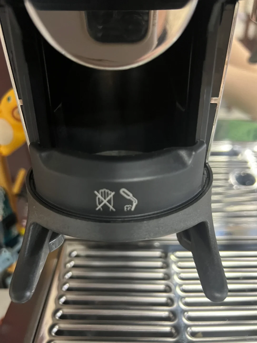 A close-up view of the portafilter cradle on an espresso machine, showing a dark gray plastic bracket with two icons: one indicating not to insert hands and another showing cleaning with a brush, positioned above a stainless steel drip tray grate.