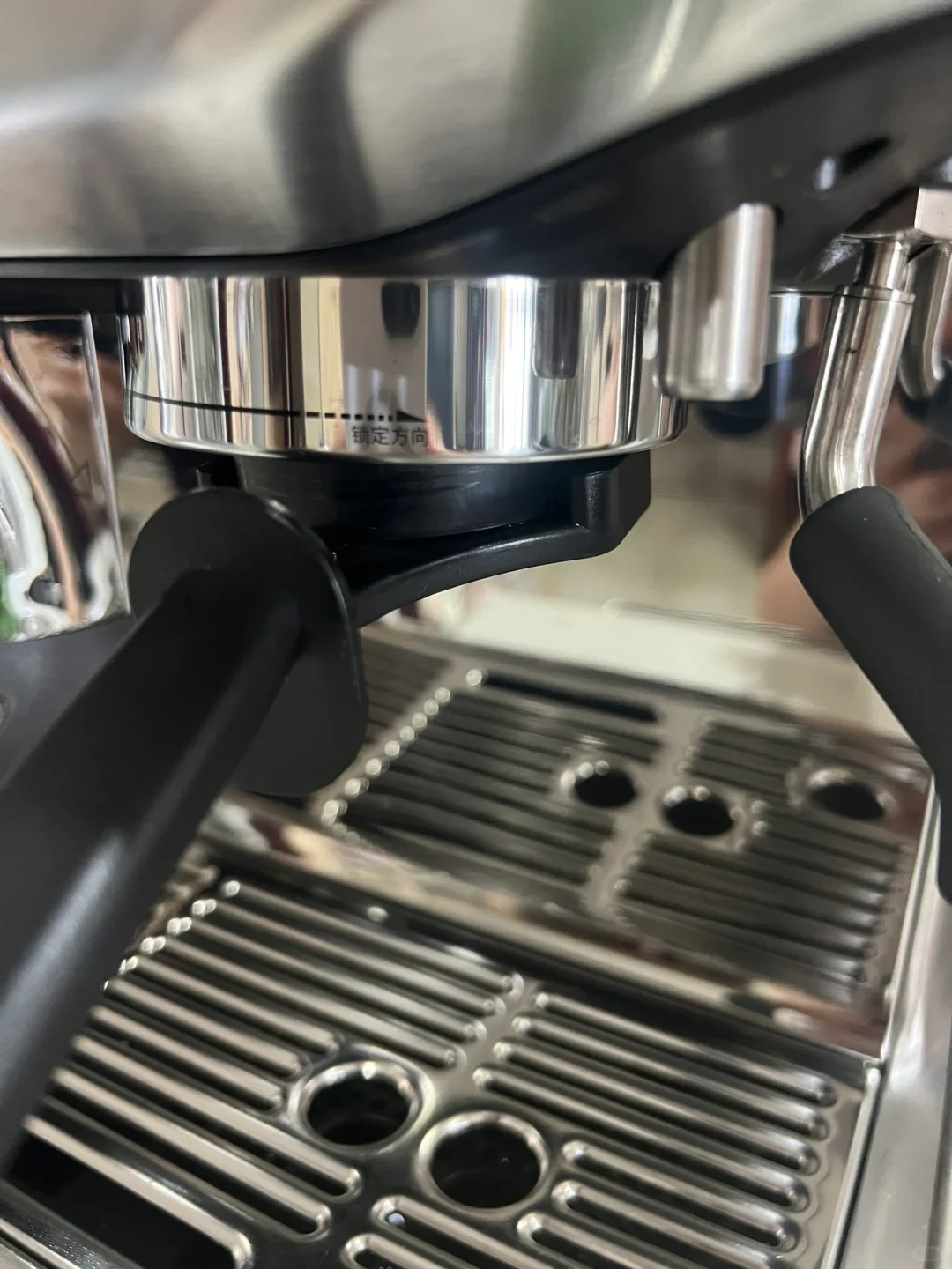 A close-up view of a stainless steel espresso machine showing the group head collar with a portafilter attached, a steam wand to the right, and the metal drip tray grate below.