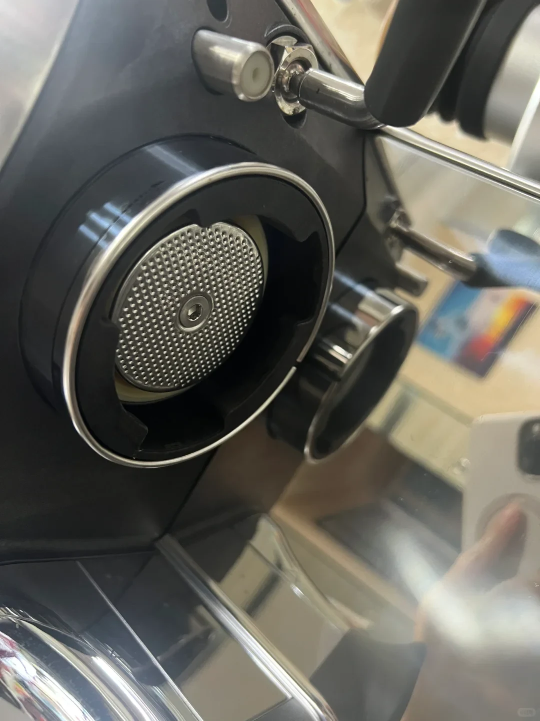 A close-up view looking up at the group head of a stainless steel espresso machine, showing the circular perforated metal shower screen secured with a central hex screw inside the black collar and chrome rim.