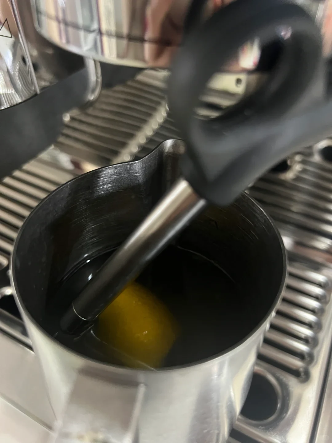 A stainless steel milk pitcher containing water and a lemon wedge sits on the drip tray of an espresso machine with the steam wand submerged inside.