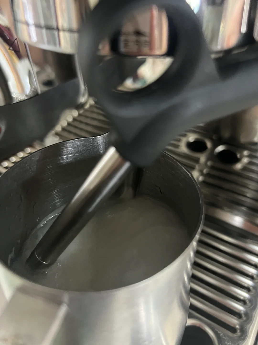 A close-up view of a stainless steel milk frothing pitcher positioned on an espresso machine's drip tray, with the black-handled steam wand nozzle inserted into liquid inside the pitcher.