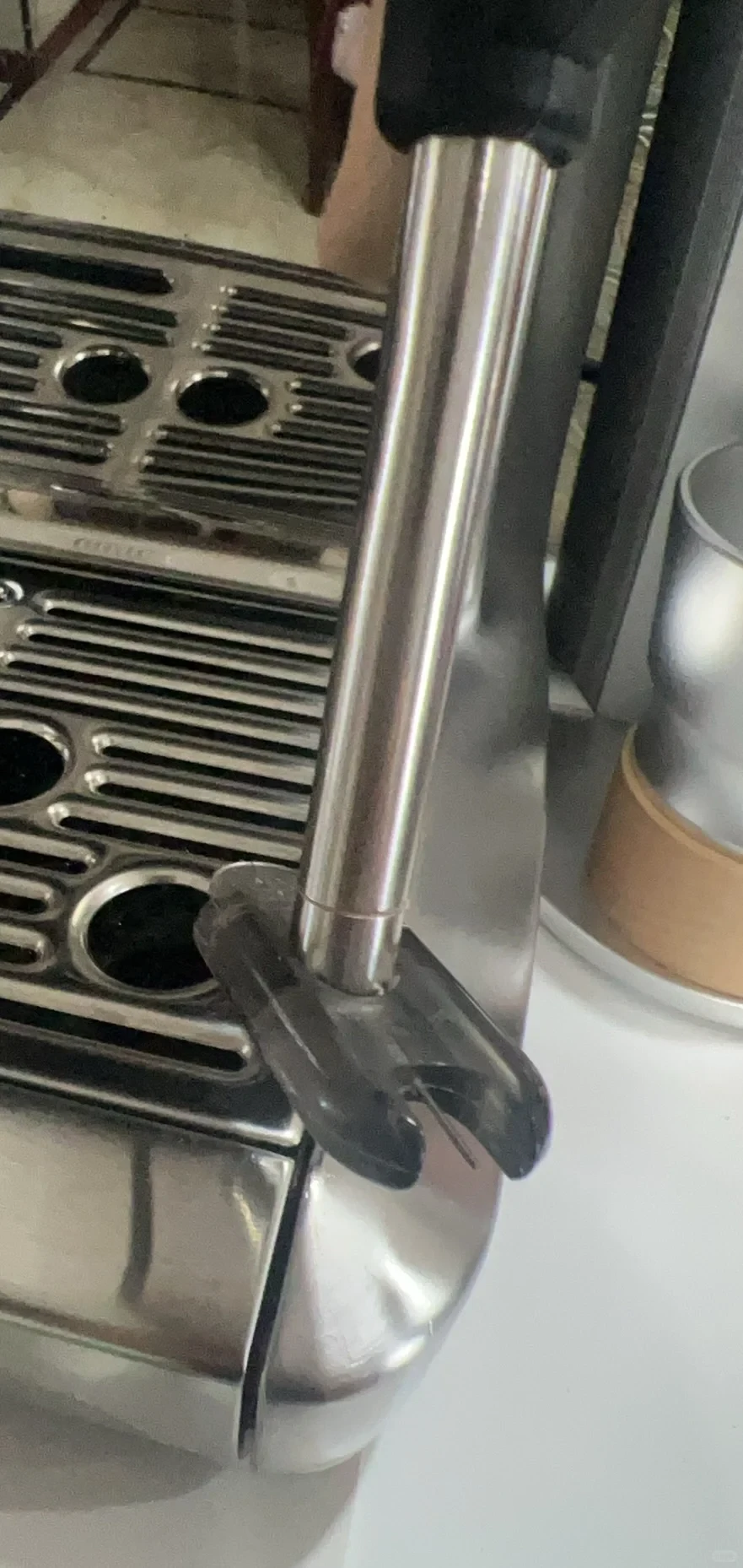 Close-up of a stainless steel espresso machine showing the drip tray grates on the left and a vertical metallic cylinder rod with a black bracket attachment at its lower end.