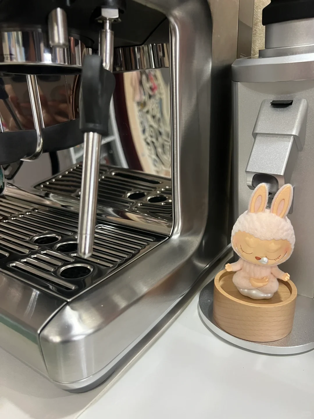 A stainless steel espresso machine with a steam wand and drip tray sits beside a silver coffee grinder, under which rests a small figurine of a sleeping rabbit character seated cross-legged on a wooden round stand.