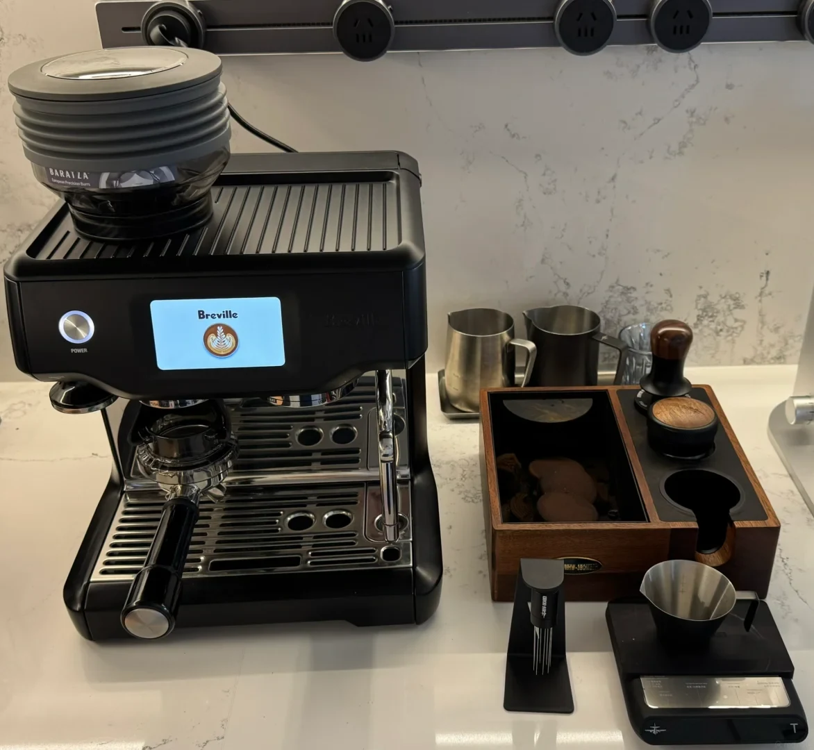 Front view of a black Breville 880 espresso machine fitted with a bellows attachment, alongside a wooden tamping station, knock box, tamper, WDT tool, digital scale, and milk pitchers.