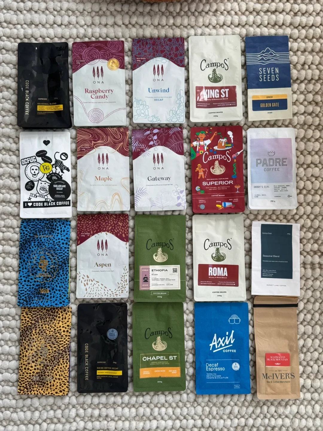 Nineteen assorted specialty coffee bean bags from Australian roasters neatly arranged in rows on a textured white rug.