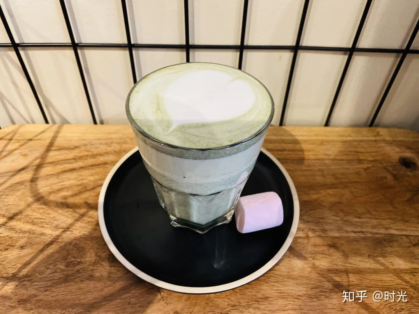 A glass of pale green, frothy coffee sits on a black saucer on a wooden table, with a pink marshmallow-like confection beside it.