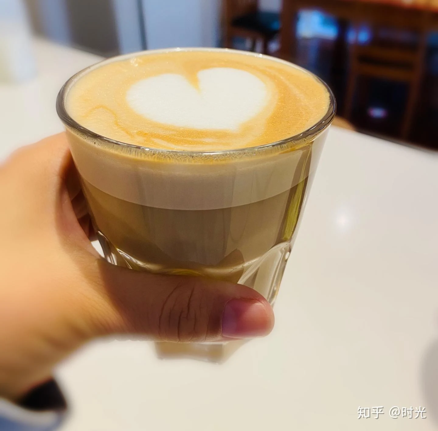 A hand holds a clear glass of coffee topped with thick pale foam and a simple white heart pattern on a white tabletop.