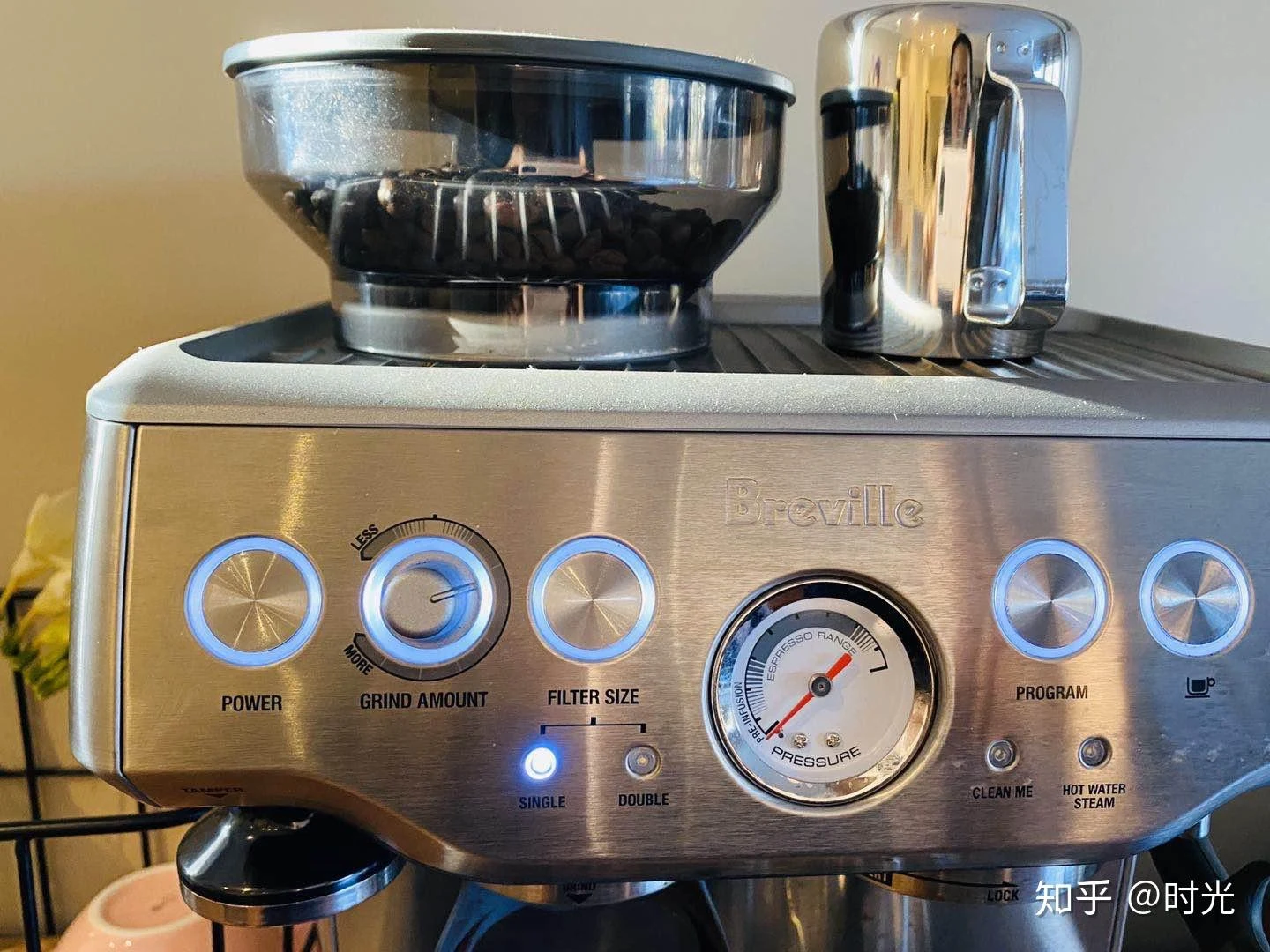 Close-up of a stainless-steel Breville espresso machine with a bean-filled grinder hopper, portafilter, blue-lit controls, and an espresso pressure gauge.
