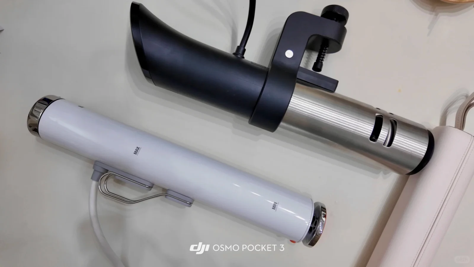 Two cylindrical sous vide immersion circulators resting diagonally on a light-colored surface. The upper cooker features a black upper handle housing, a black mounting clamp with an adjustment knob, and a brushed stainless steel lower cylinder. The lower cooker is slender and white with chrome end caps, a wire clip, and marked MAX and MIN water level indicators.