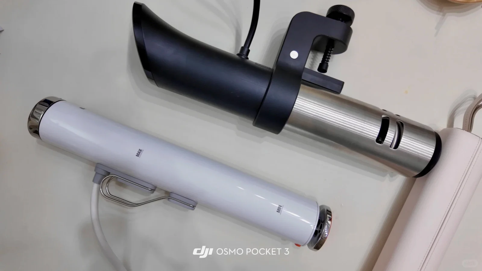 Top-down view of two immersion circulators lying side by side on a flat surface. The top device features a black contoured upper body, an attached mounting clamp with a screw knob, and a brushed stainless steel lower cylinder. The bottom device is a slender white cylinder featuring a wire clip and printed 'MAX' and 'MIN' fill lines. A 'DJI OSMO POCKET 3' watermark appears at the bottom center.