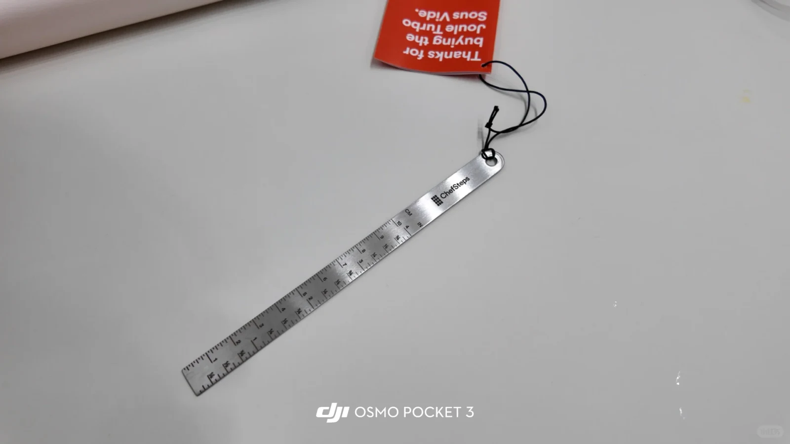 A brushed metal culinary ruler labeled ChefSteps with measurement markings in centimeters and inches, attached by a black cord to an upside-down red tag reading "Thanks for buying the Joule Turbo Sous Vide.", resting diagonally on a white surface.