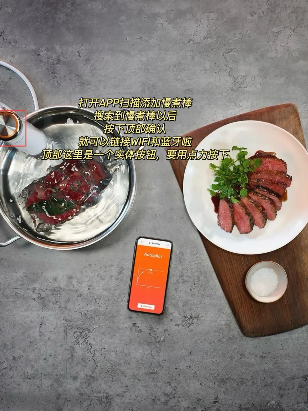 Top-down view of a sous vide cooking setup on a gray countertop. On the left, a stainless steel pot holds vacuum-bagged meat submerged in water with a sous vide immersion circulator clamped to the side, featuring a red box around its top button. In the center, a smartphone displays an orange app screen labeled Autopilot with a temperature curve. On the right, a wooden board holds a white plate of sliced medium-rare steak garnished with watercress alongside a small bowl of flaky salt.