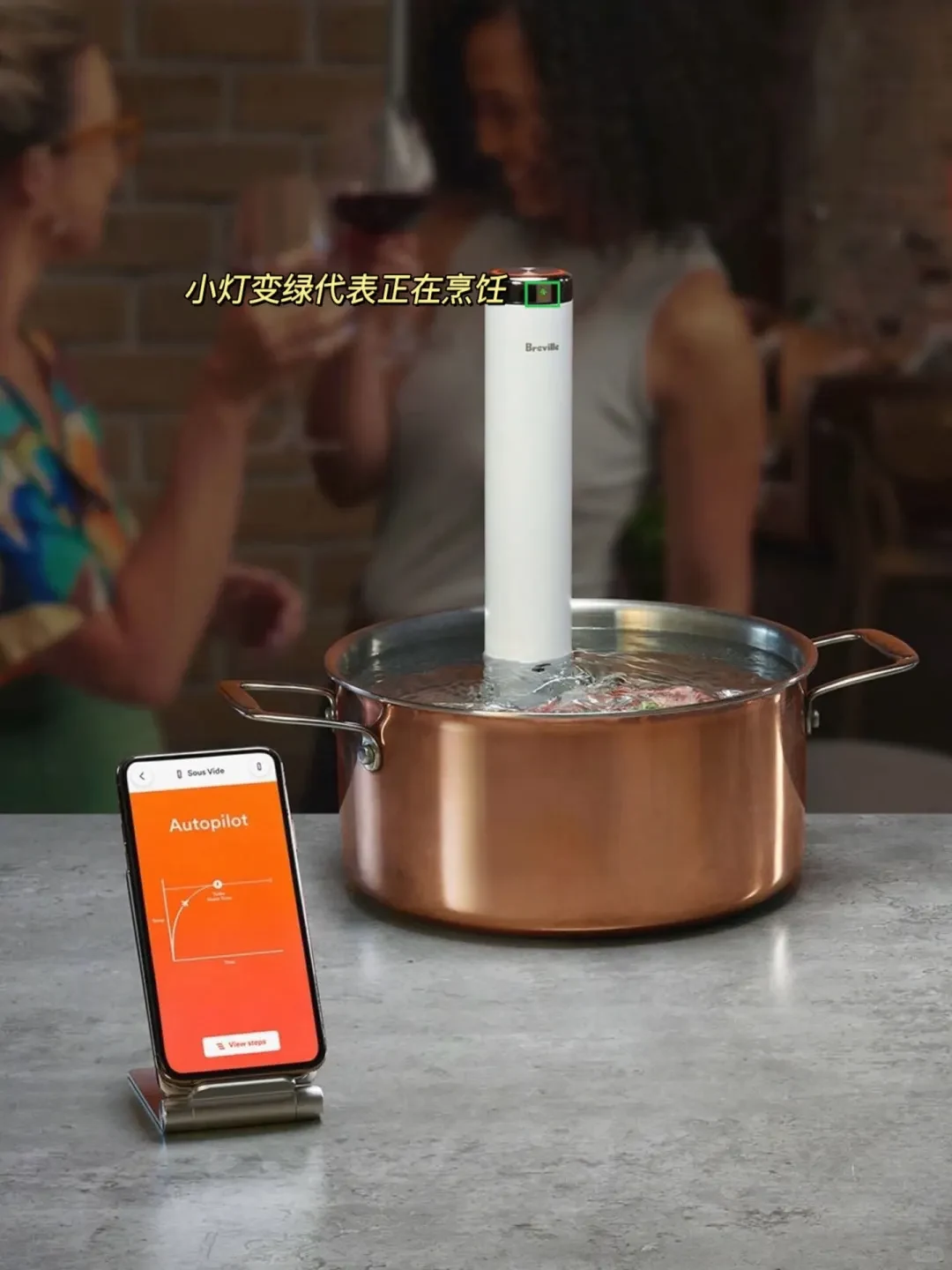 A white Breville sous vide immersion circulator operating inside a copper pot of water on a kitchen counter, beside a smartphone displaying a sous vide app on a stand.