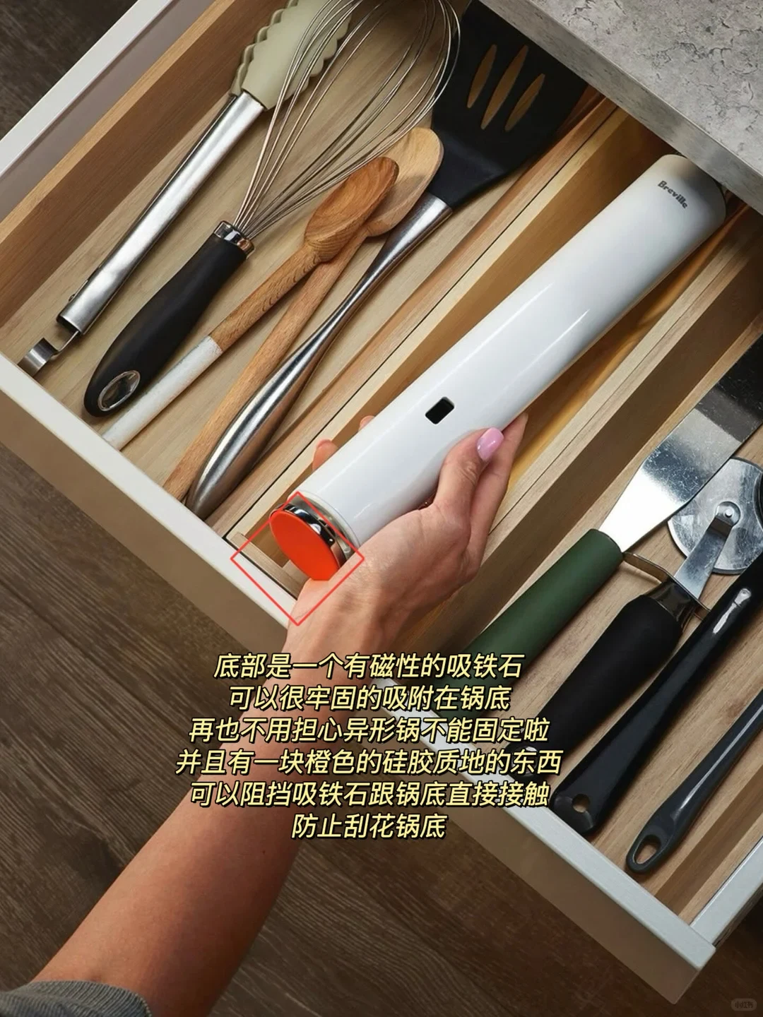 A hand holds a white Breville Joule immersion circulator above a wooden kitchen drawer filled with neatly organized utensils. A red outlined square highlights the orange silicone base attached to the bottom of the sous vide device.