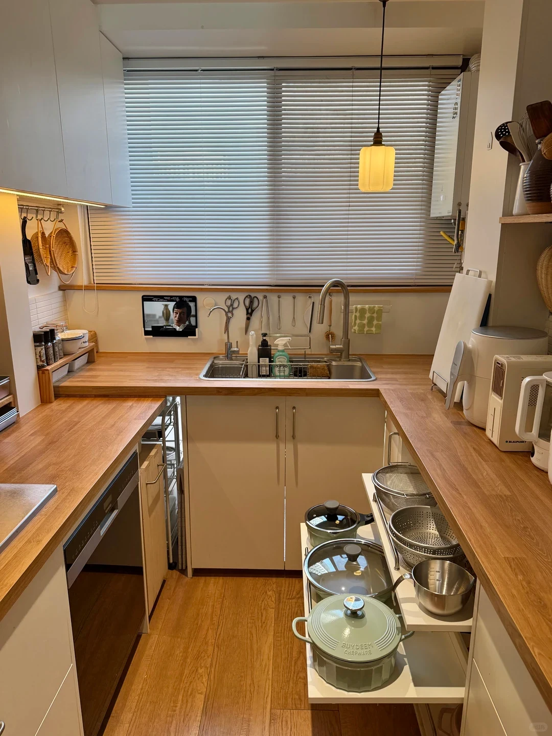 A cozy, compact U-shaped kitchen featuring warm wood countertops and white cabinetry. In the center, a stainless steel sink sits beneath a window with closed horizontal blinds and a warm pendant lamp. A small tablet screen is mounted on the wall below the window. The lower right cabinet has an extended pull-out organizer holding various pots, glass lids, and stainless steel colanders.