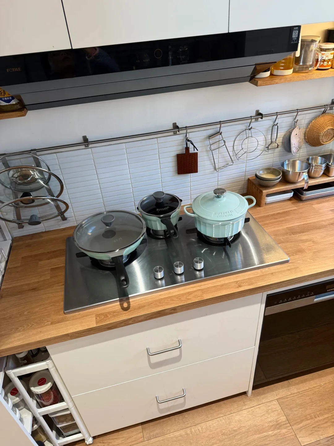 A modern kitchen countertop with a built-in stainless steel three-burner stovetop holding three mint-green Buydeem cookware pieces: a frying pan, a small saucepan, and a lidded Dutch oven. White subway tiles and a hanging utensil rail line the wall beneath a black range hood.