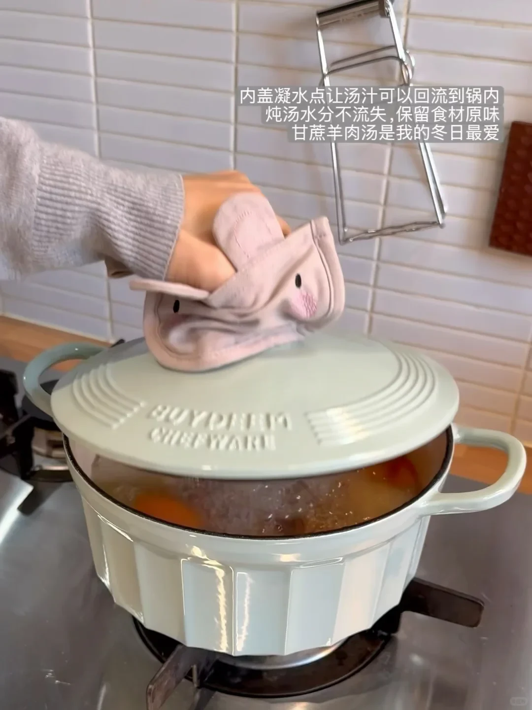 A hand using an oven mitt lifts the lid off a light green Dutch oven on a stove, revealing simmering soup inside.