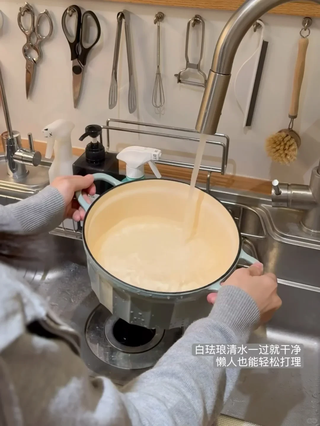 A person holds a light-blue enamel Dutch oven under a running kitchen faucet in a sink, rinsing out its pale interior.