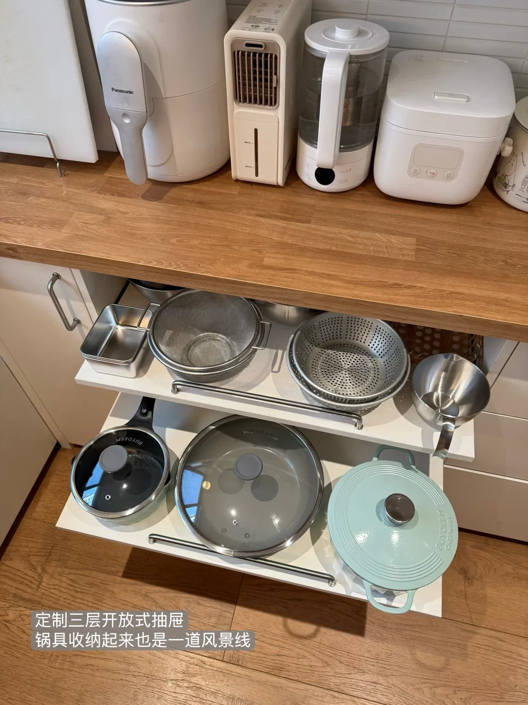 A kitchen scene showing a wooden countertop with white small appliances, and open pull-out cabinet shelves beneath storing various stainless steel strainers, cooking pans with glass lids, and a pastel blue enameled pot.