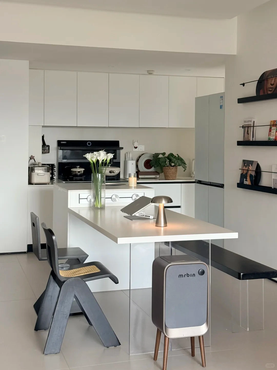 A modern open-concept white kitchen and dining space with an integrated island dining table supported by a clear acrylic base, black geometric dining chairs, a small trash bin on wooden legs labeled 'mrbin', and white cabinetry.