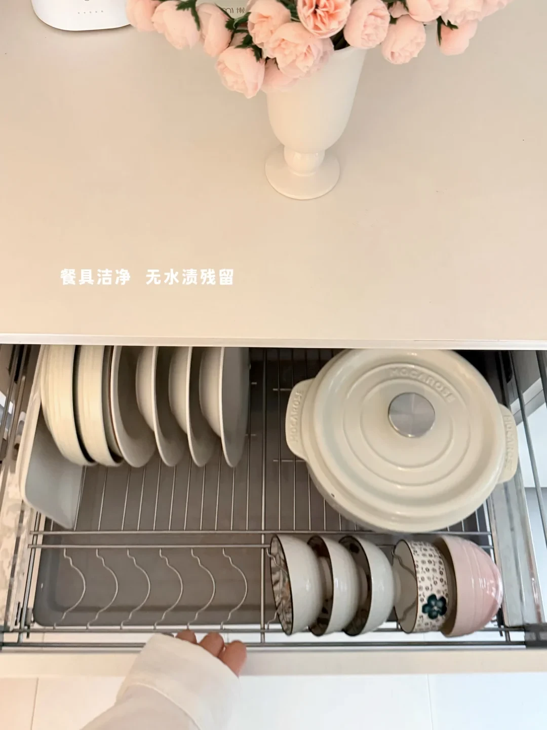 A pull-out kitchen cabinet dish rack drawer shown from above, loaded with clean plates, small bowls, and a round white lidded Dutch oven beneath a white countertop with a vase of pink flowers.